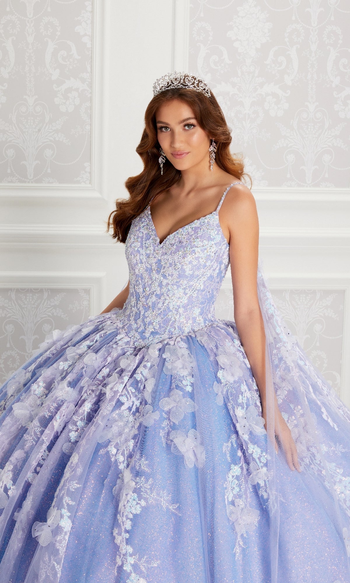 Woman wearing the Quinceanera Dress PR22143 By Princesa, a sparkling lavender gown with floral details and beaded corset bodice, paired with a tiara and drop earrings, standing against an ornate white background.